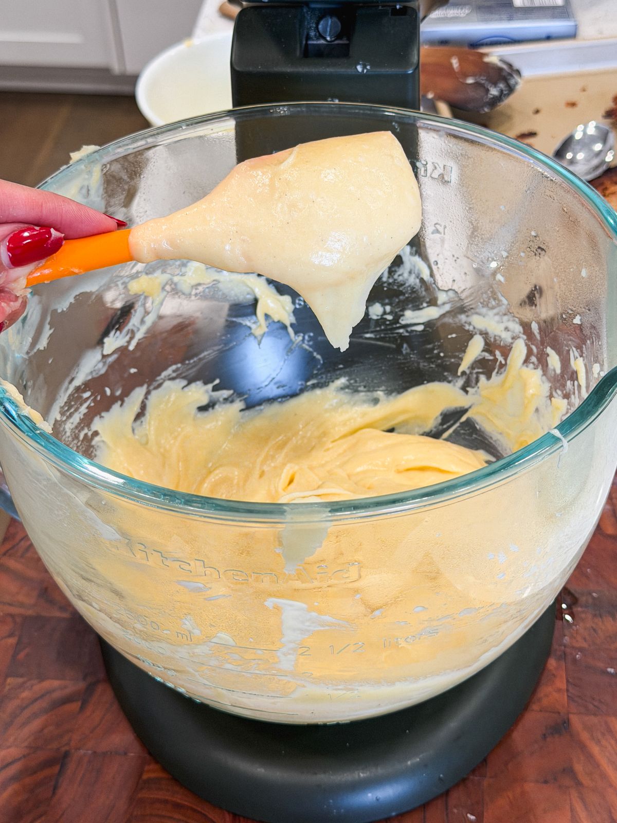 A hand holding a spatula with thick, choux pastry over a glass mixing bowl attached to a stand mixer, sitting on a wooden countertop. The bowl contains more batter, and kitchen items are visible in the background. The choux pastry has fallen off the spatula leaving behind a rough v shape