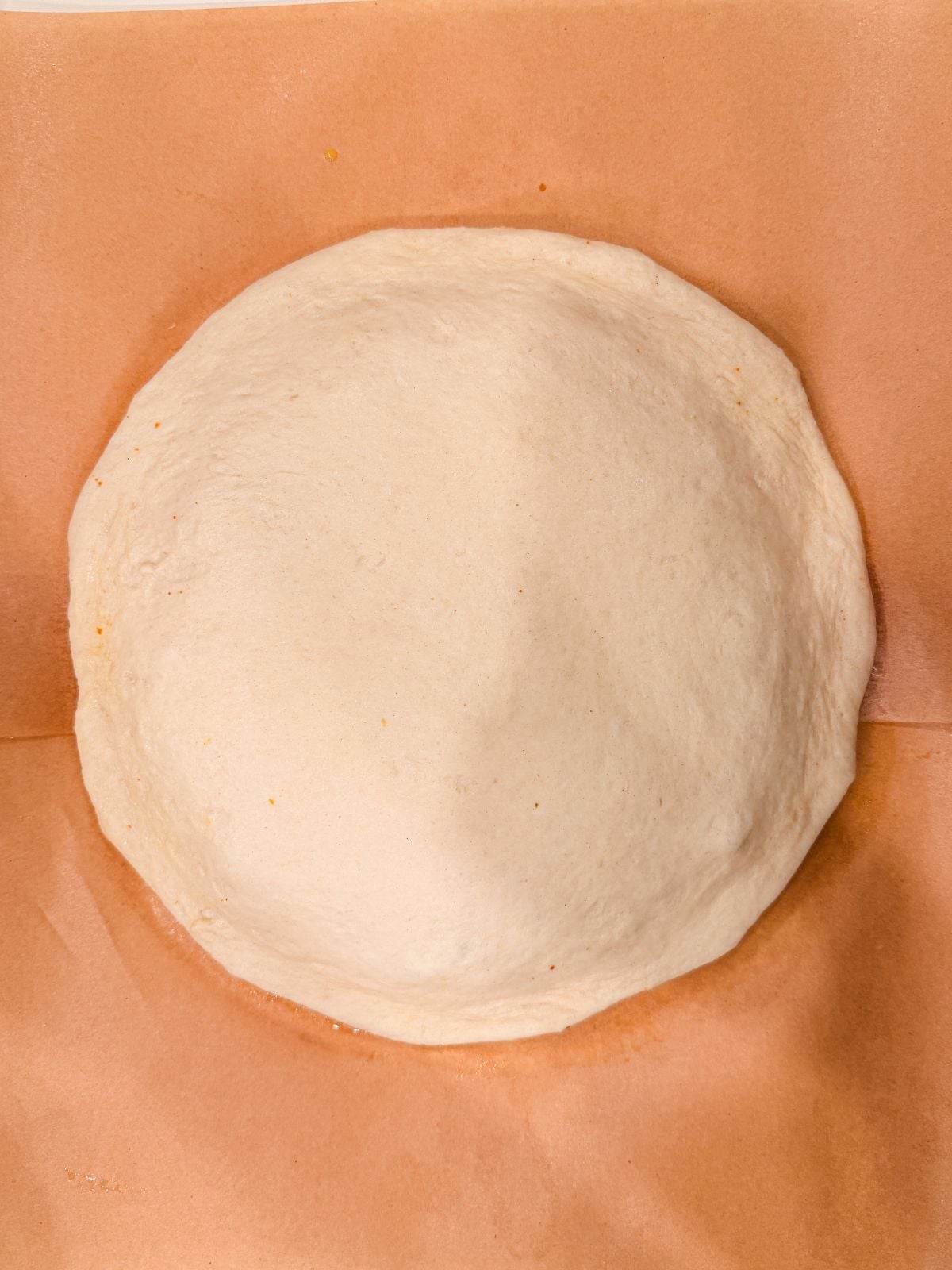 A round, uncooked pizza dough is placed on a sheet of brown parchment paper. The dough is light in color and has a slightly raised edge, ready for toppings.
