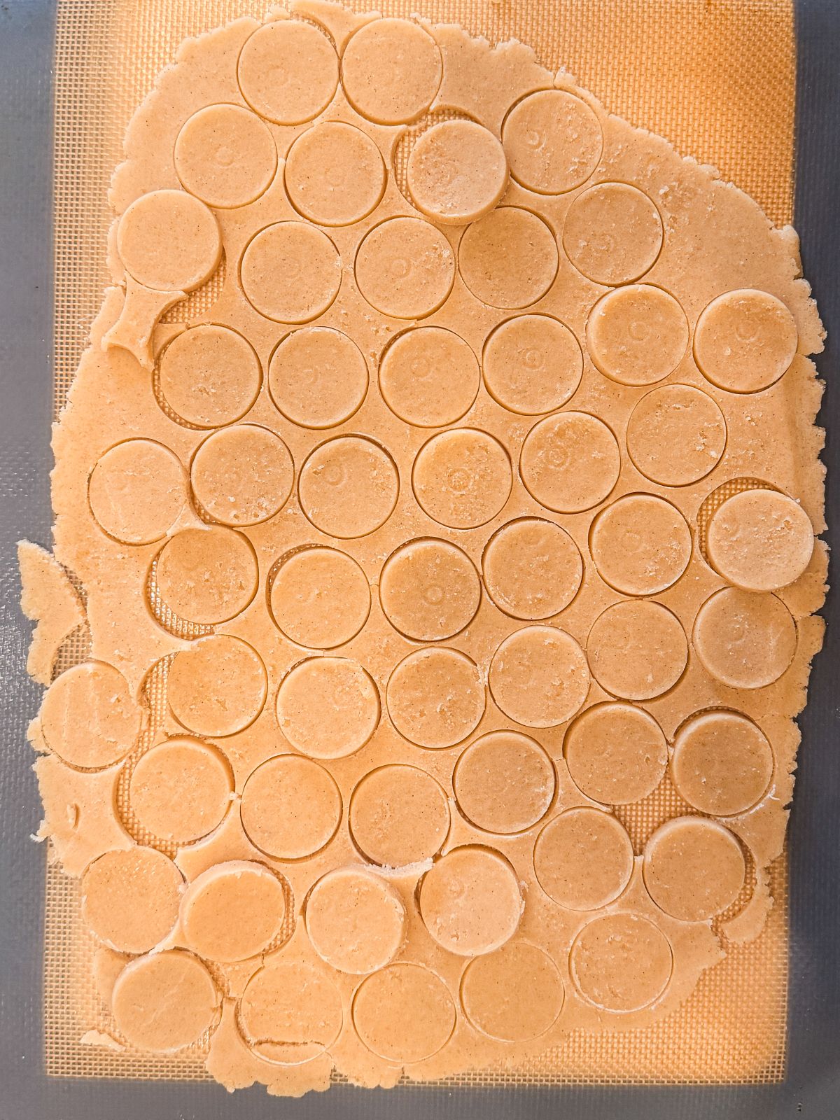 Circular craquelin shapes cut out on a rolled sheet of craquelin, arranged closely together on a silicone baking mat, with some dough scraps remaining around the edges.