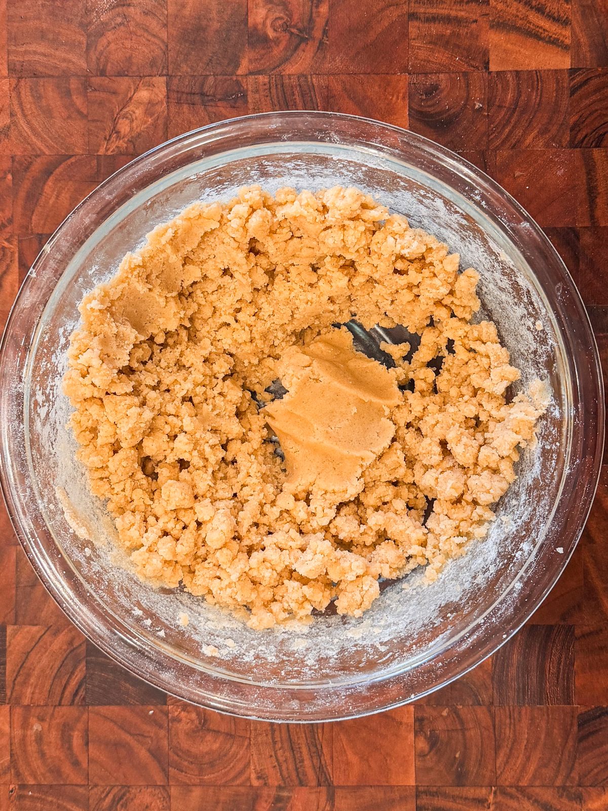A glass bowl on a wooden surface contains crumbly, craquelin dough, with some dough pressed together in the center.