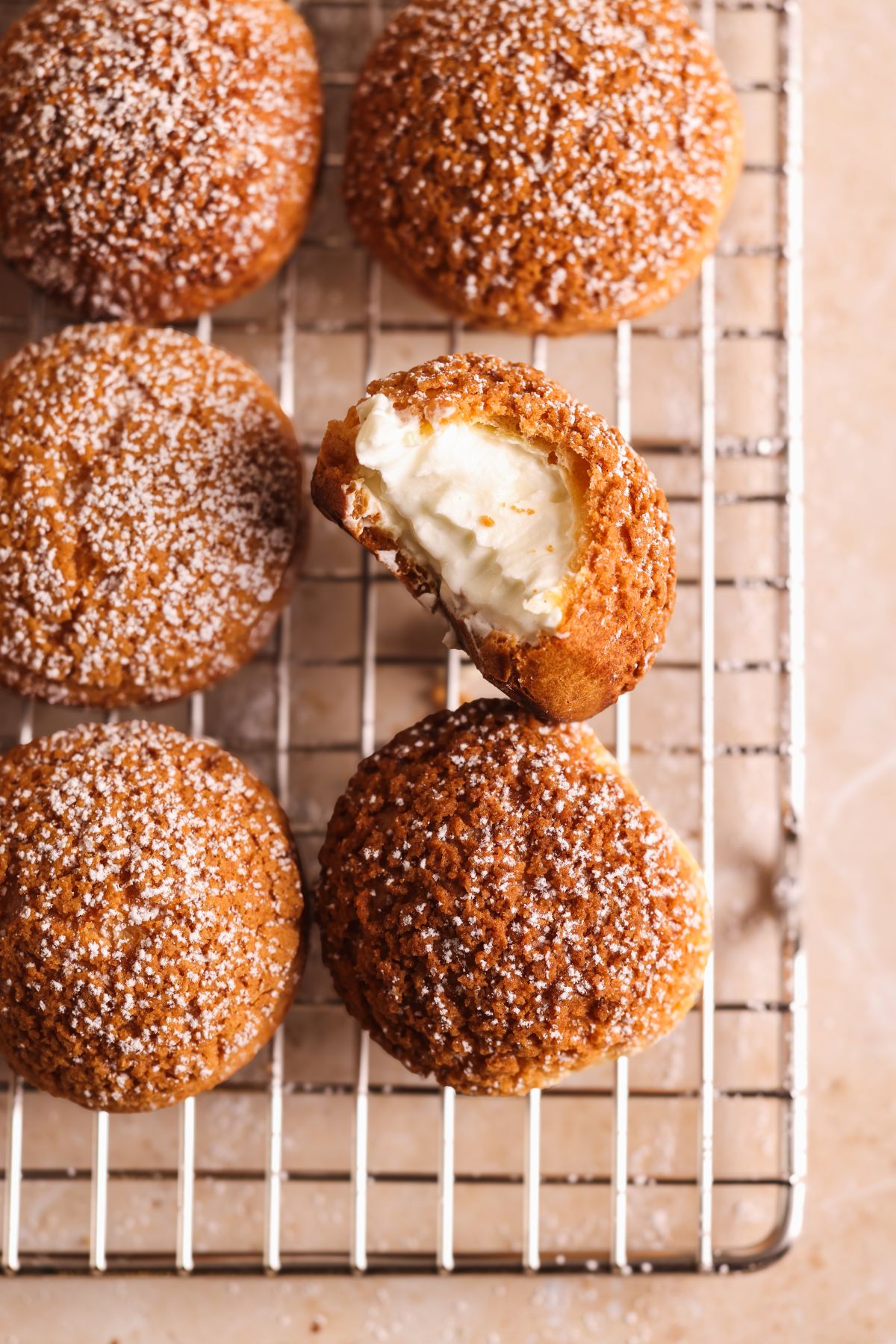 Six round cream puffs dusted with powdered sugar rest on a cooling rack, with one puff centered and bitten into, revealing its creamy filling. The background surface is light beige.