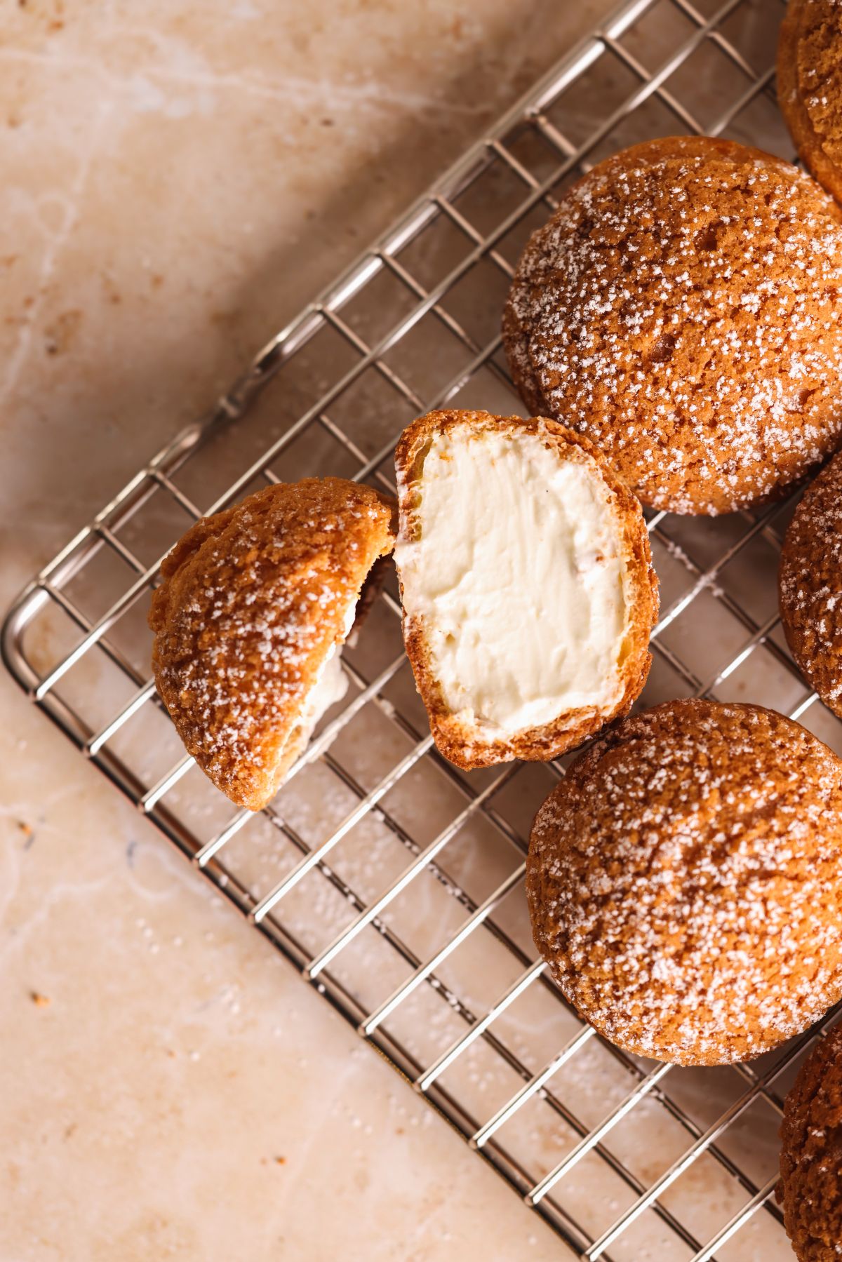 A wire rack holds round cream puffs dusted with powdered sugar; one is cut open to reveal a rich, creamy filling inside.