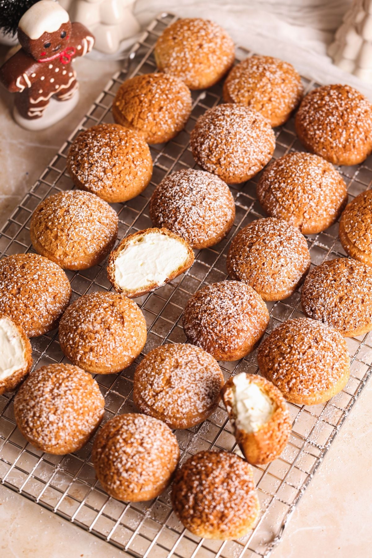Cream puffs dusted with powdered sugar are arranged on a wire cooling rack. Two puffs are cut open, revealing a creamy filling inside. A gingerbread cookie is visible in the top left corner.