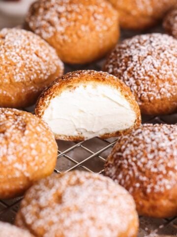 Cream puffs topped with powdered sugar are arranged on a cooling rack. One cream puff is cut in half, revealing a generous, creamy white filling inside.