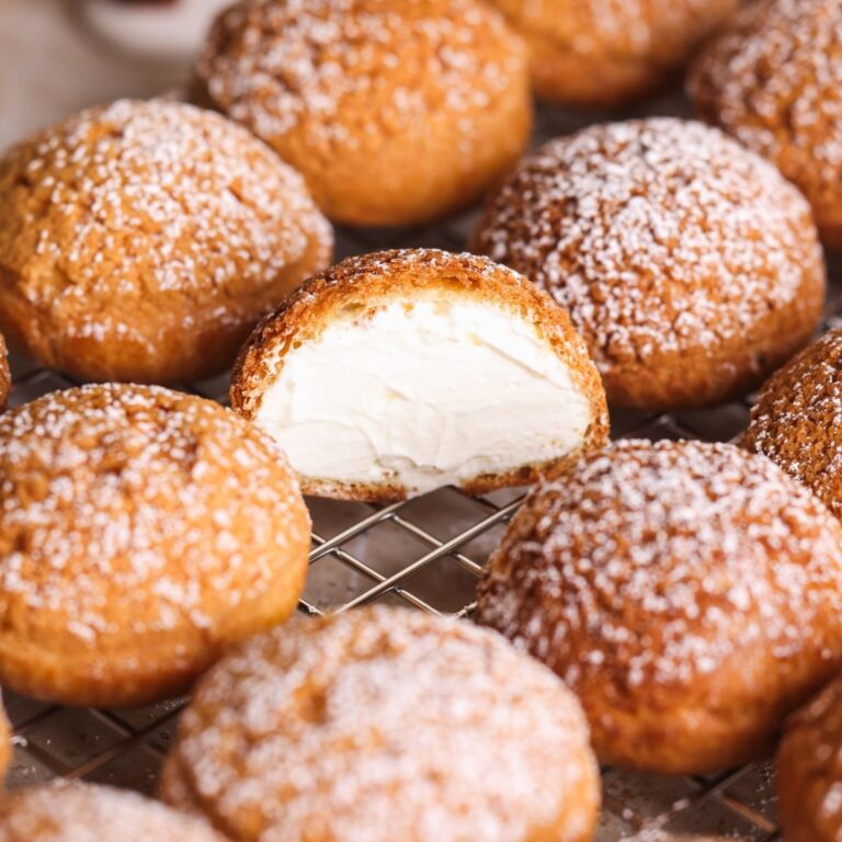 Cream puffs topped with powdered sugar are arranged on a cooling rack. One cream puff is cut in half, revealing a generous, creamy white filling inside.
