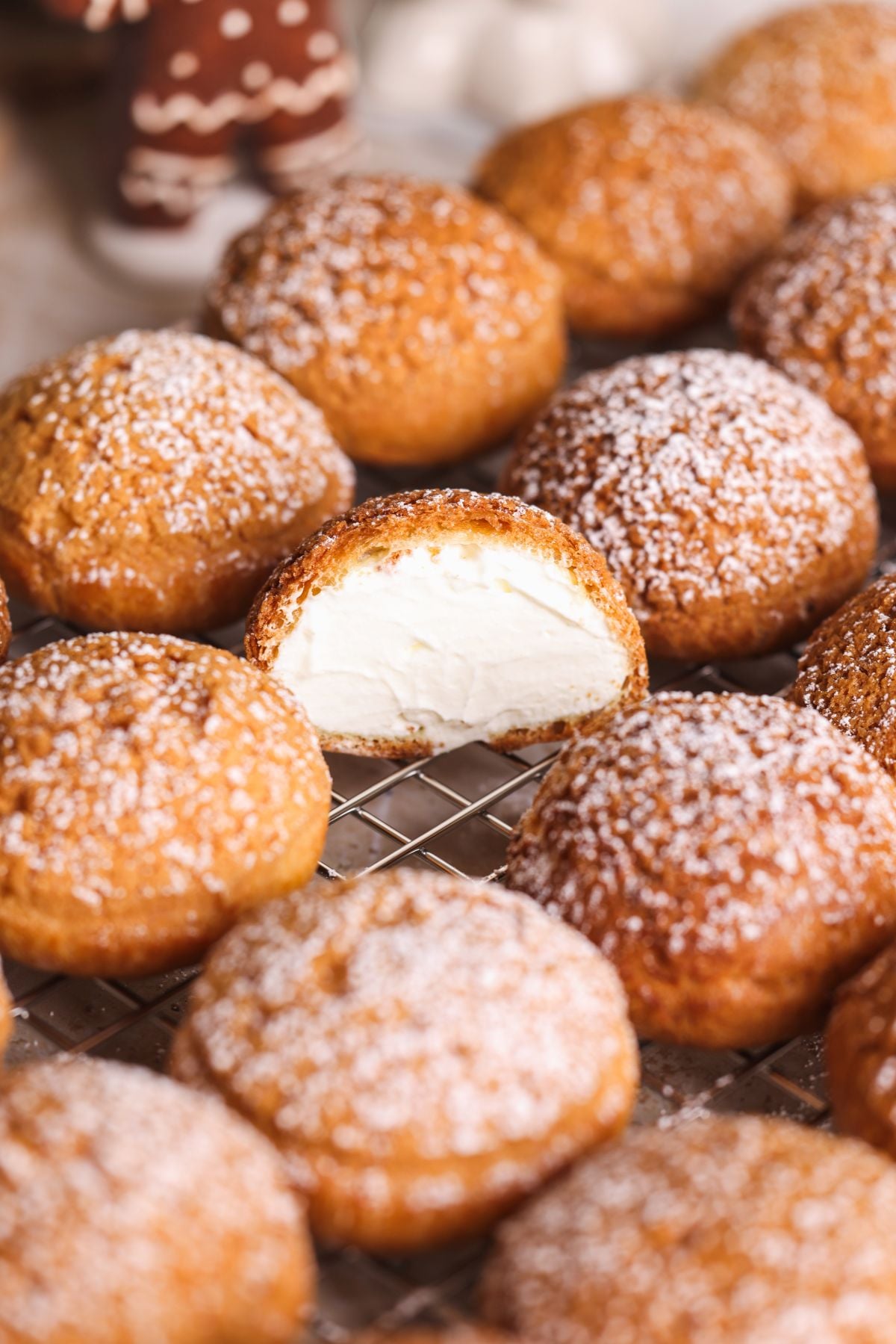 Cream puffs dusted with powdered sugar are arranged on a cooling rack. One cream puff is cut open, revealing a smooth, white cream filling inside.