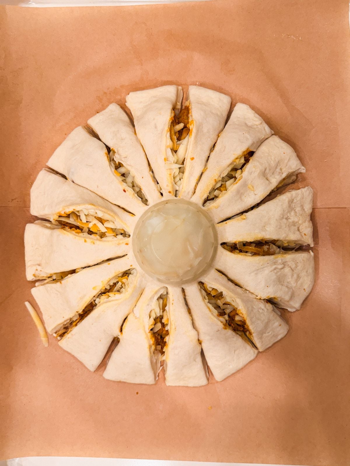 A round ring of dough with shredded cheese and filling visible in each section, sliced into segments around a small bowl placed in the center, all on a sheet of parchment paper.