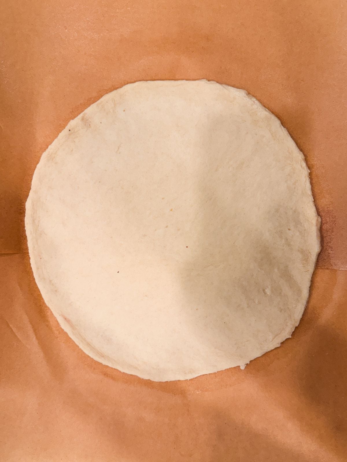 A round, uncooked pizza dough sits on a sheet of brown parchment paper, viewed from above.