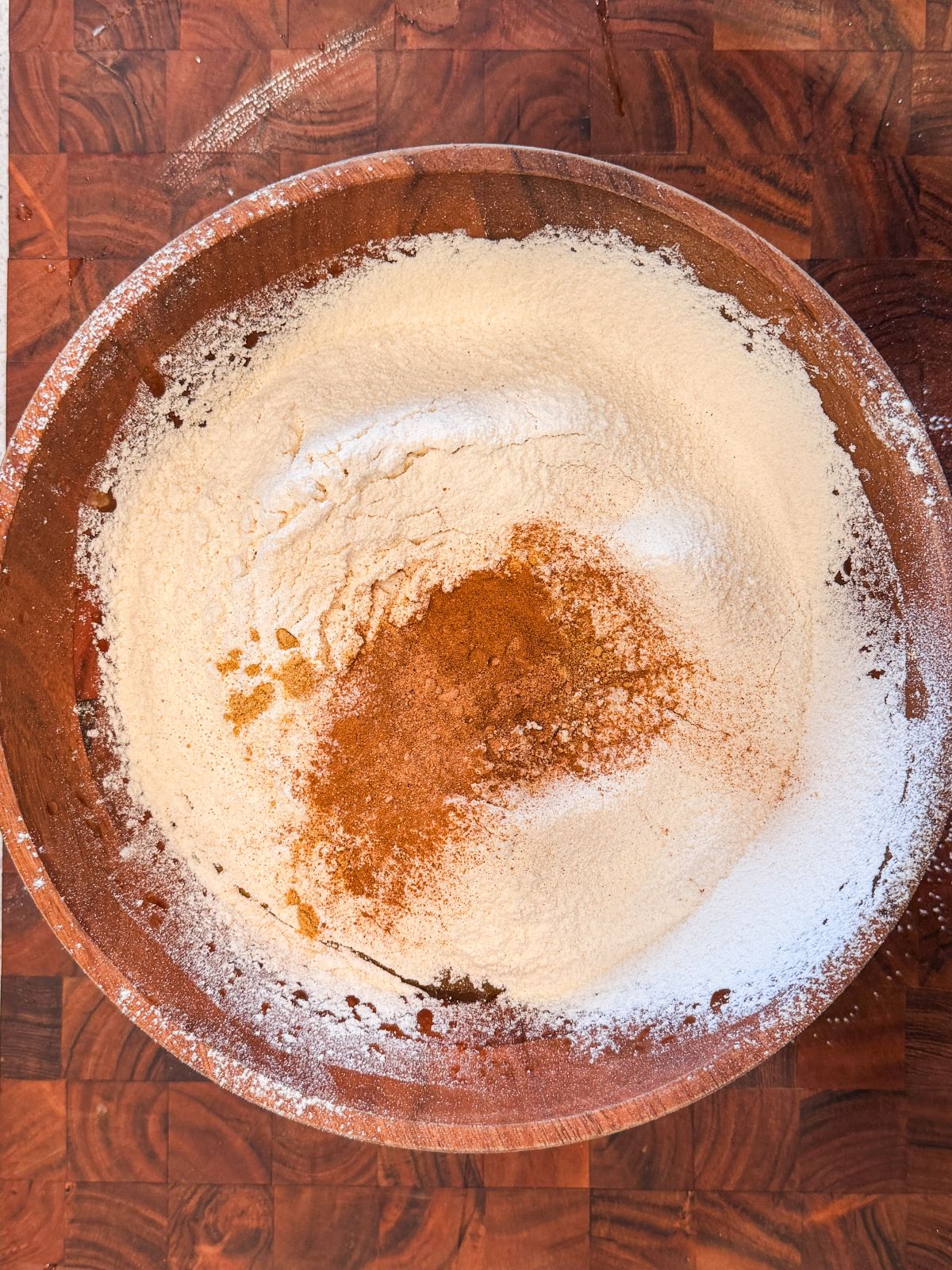 A wooden bowl filled with flour and a heap of ground cinnamon sits on a dark wooden countertop, ready for baking preparation.