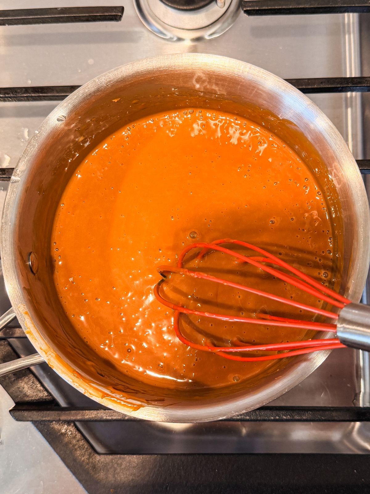 A pot of dulce de leche cream being stirred with a red whisk on a stovetop. The sauce appears smooth and creamy, with small bubbles on the surface.