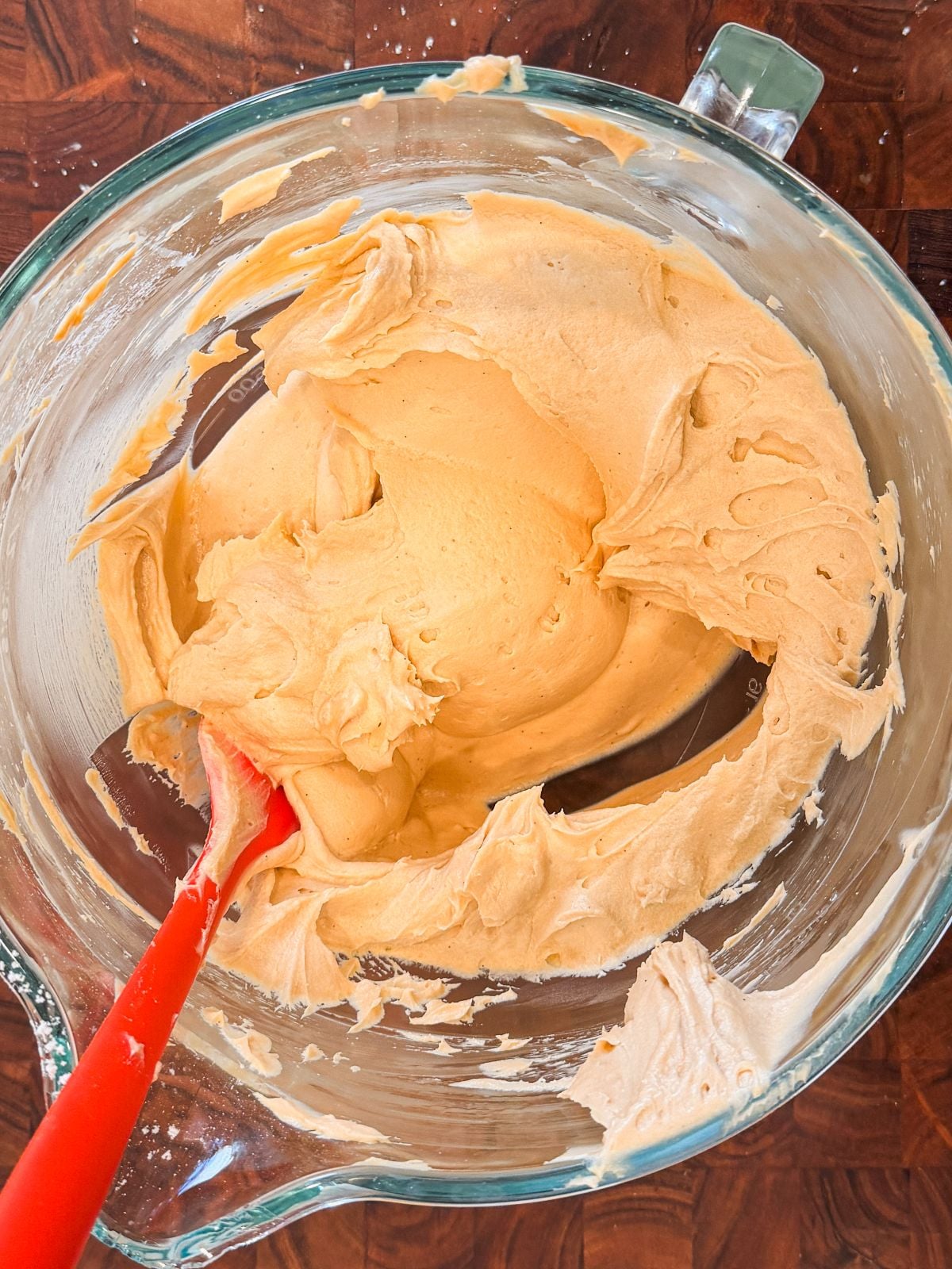 A glass mixing bowl filled with creamy, light frosting being mixed with a red spatula, placed on a wooden surface.