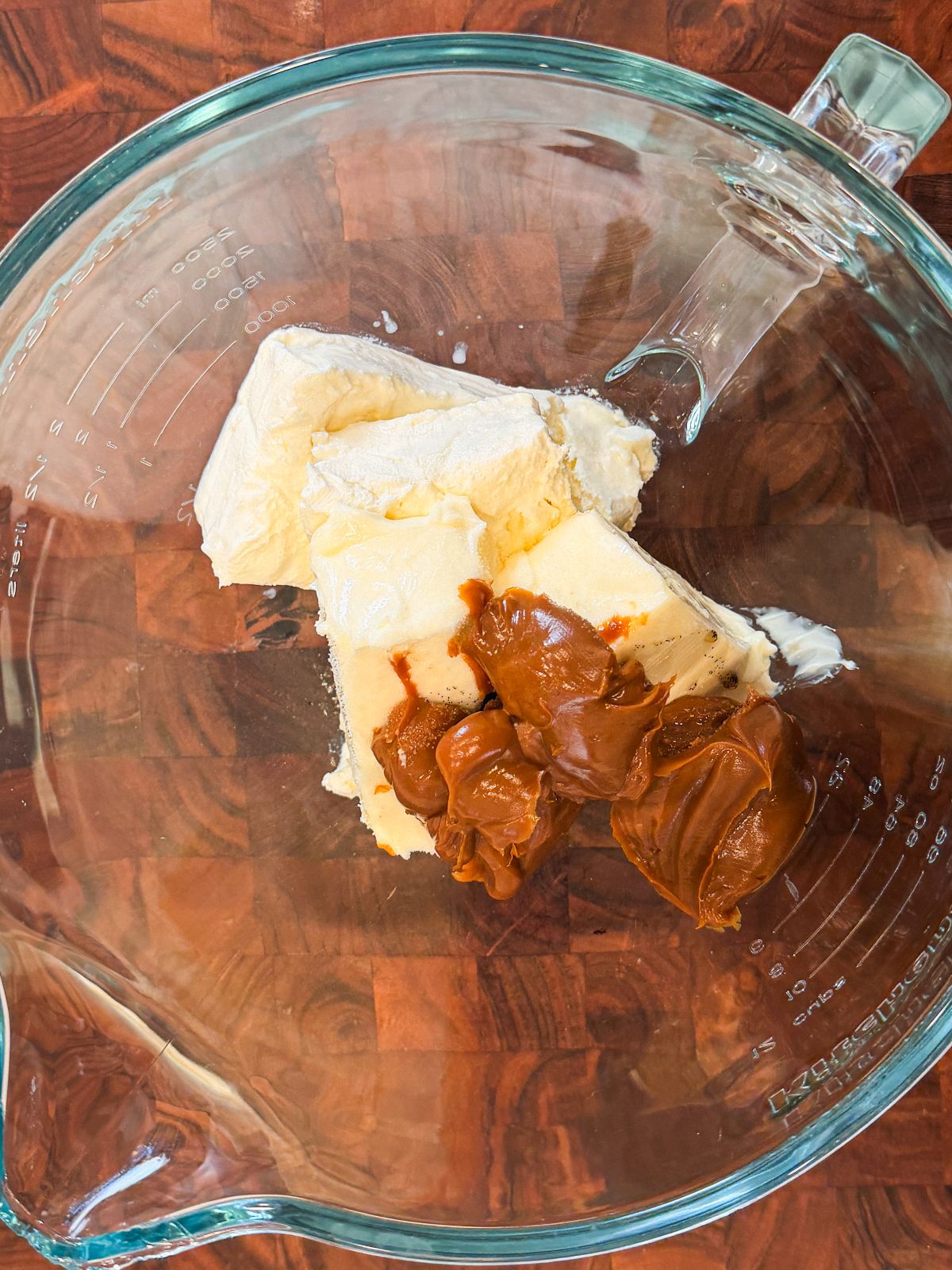 A glass mixing bowl containing cream cheese and a few dollops of dulce de leche, placed on a wooden surface.