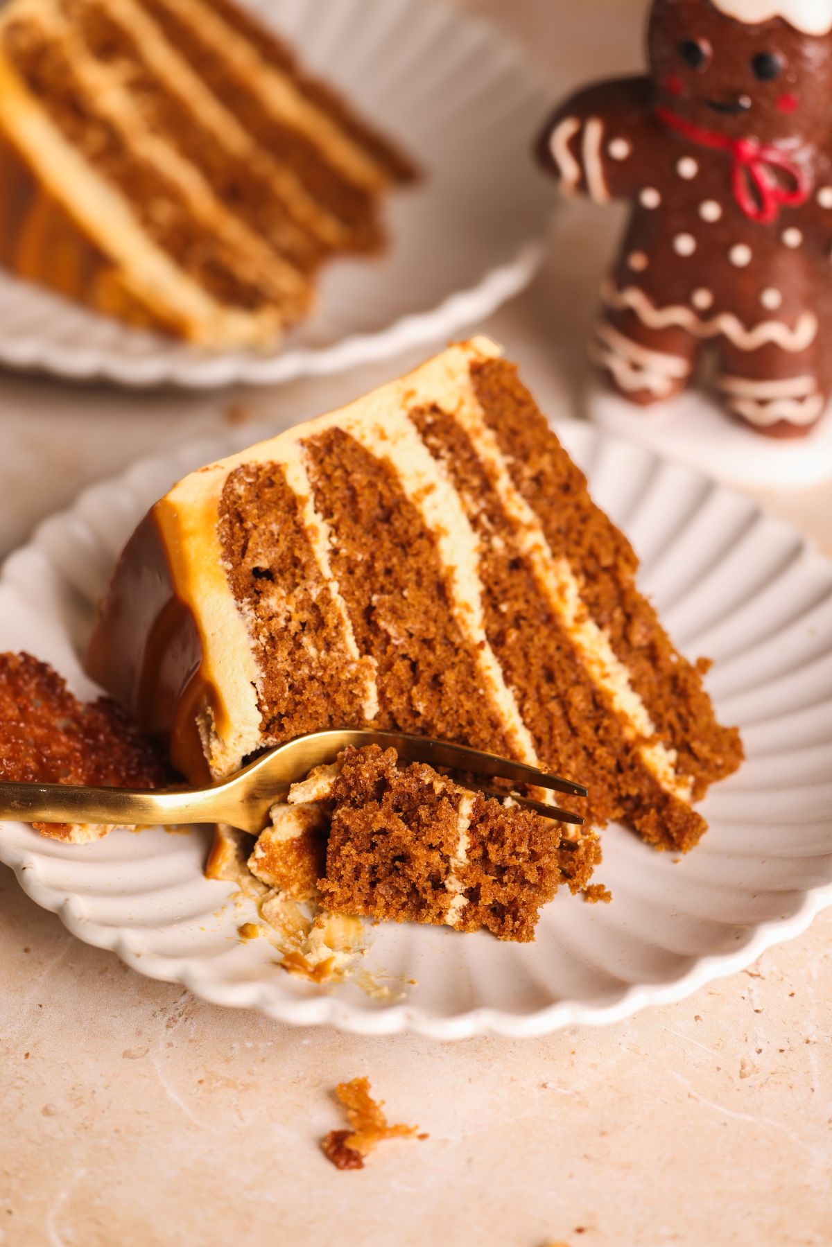 A slice of layered gingerbread cake with cream cheese frosting on a white plate, a fork taking a bite, and a gingerbread man cookie in the background.