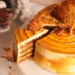 A dulce de leche glazed gingerbread layer cake with a slice being lifted out, revealing multiple layers of cake and filling, topped with ginger snaps, on a white cake stand.