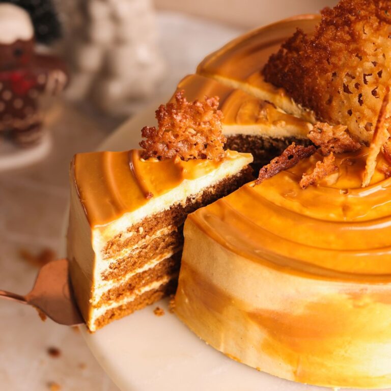 A dulce de leche glazed gingerbread layer cake with a slice being lifted out, revealing multiple layers of cake and filling, topped with ginger snaps, on a white cake stand.