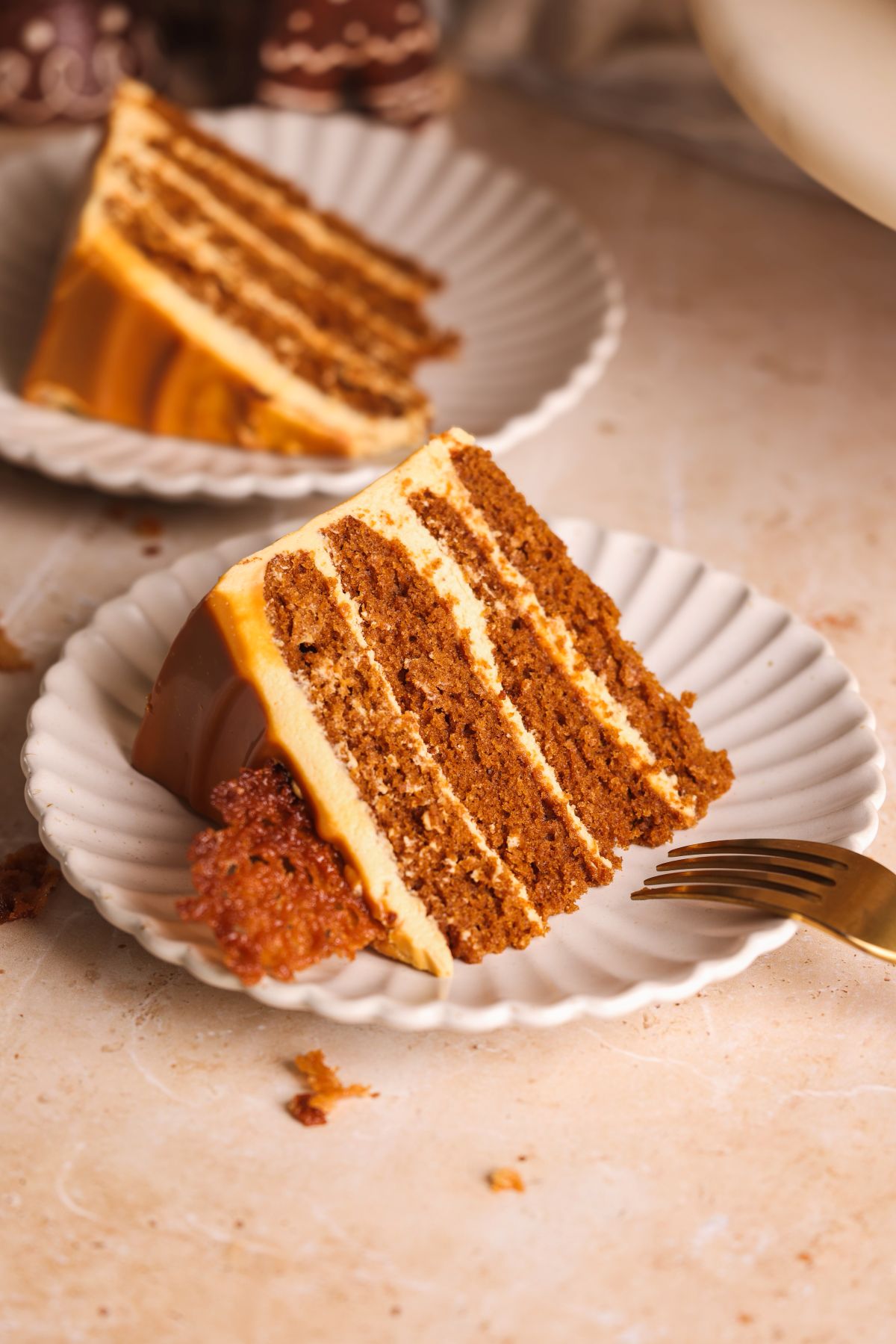 A slice of layered gingerbread cake with dulce de leche frosting sits on a scalloped white plate, with a gold fork beside it. Another plate with cake is in the background on a light-colored surface.
