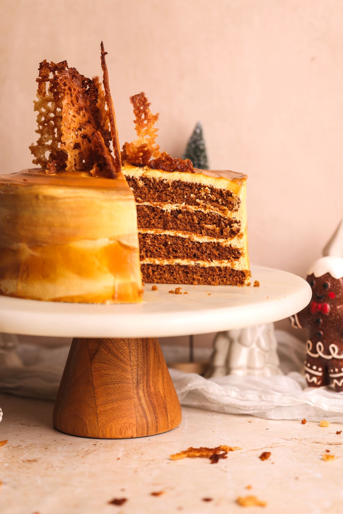 A layered cake with caramel-colored frosting and decorative ginger snaps sits on a wooden cake stand. A slice is missing, revealing the cakes layers. There are gingerbread decorations and crumbs nearby.