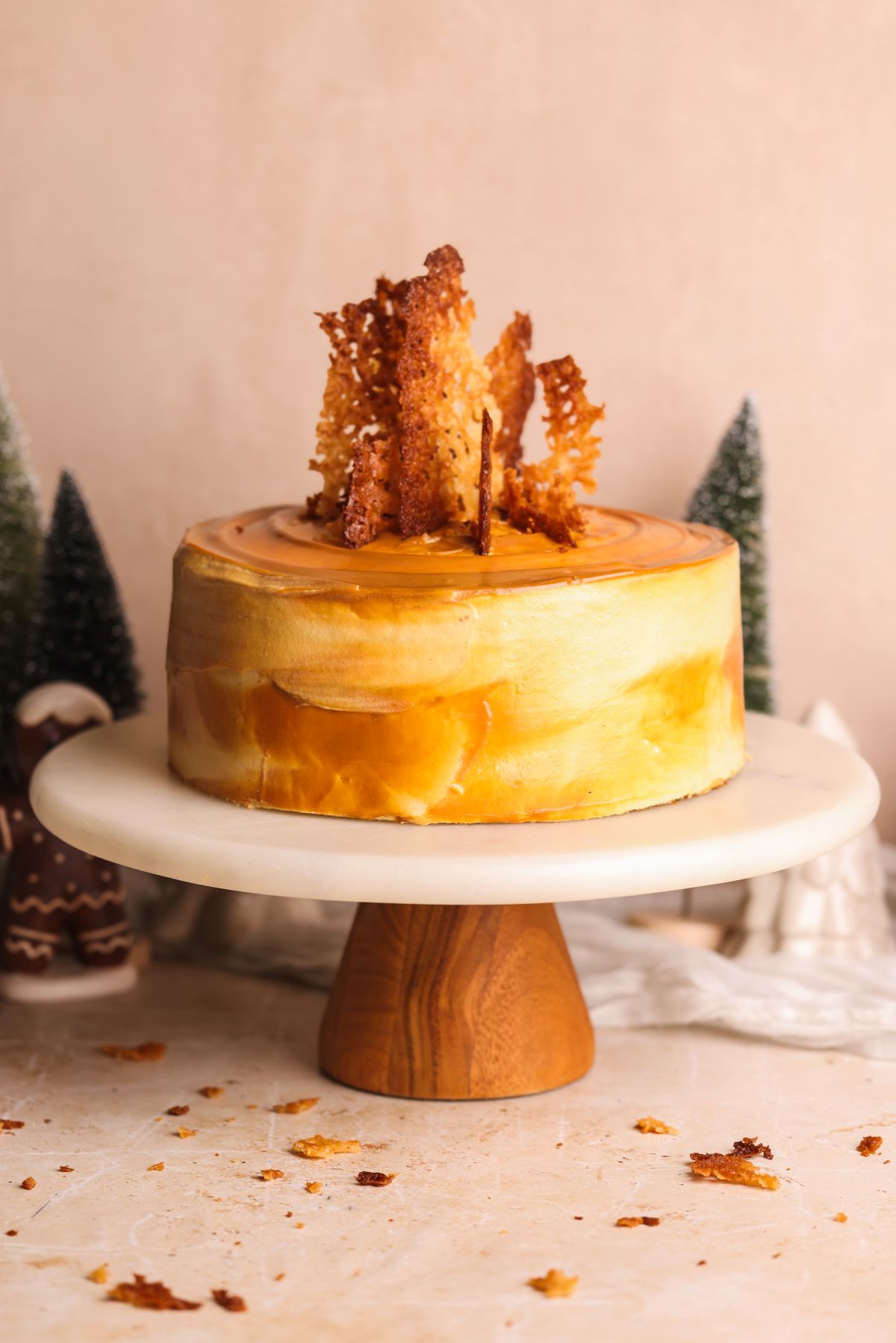A caramel-colored frosted gingerbread cake with crisp, golden ginger snaps on top sits on a wooden cake stand. Small decorative trees and gingerbread figures are in the background. Crumbs are scattered on the surface.