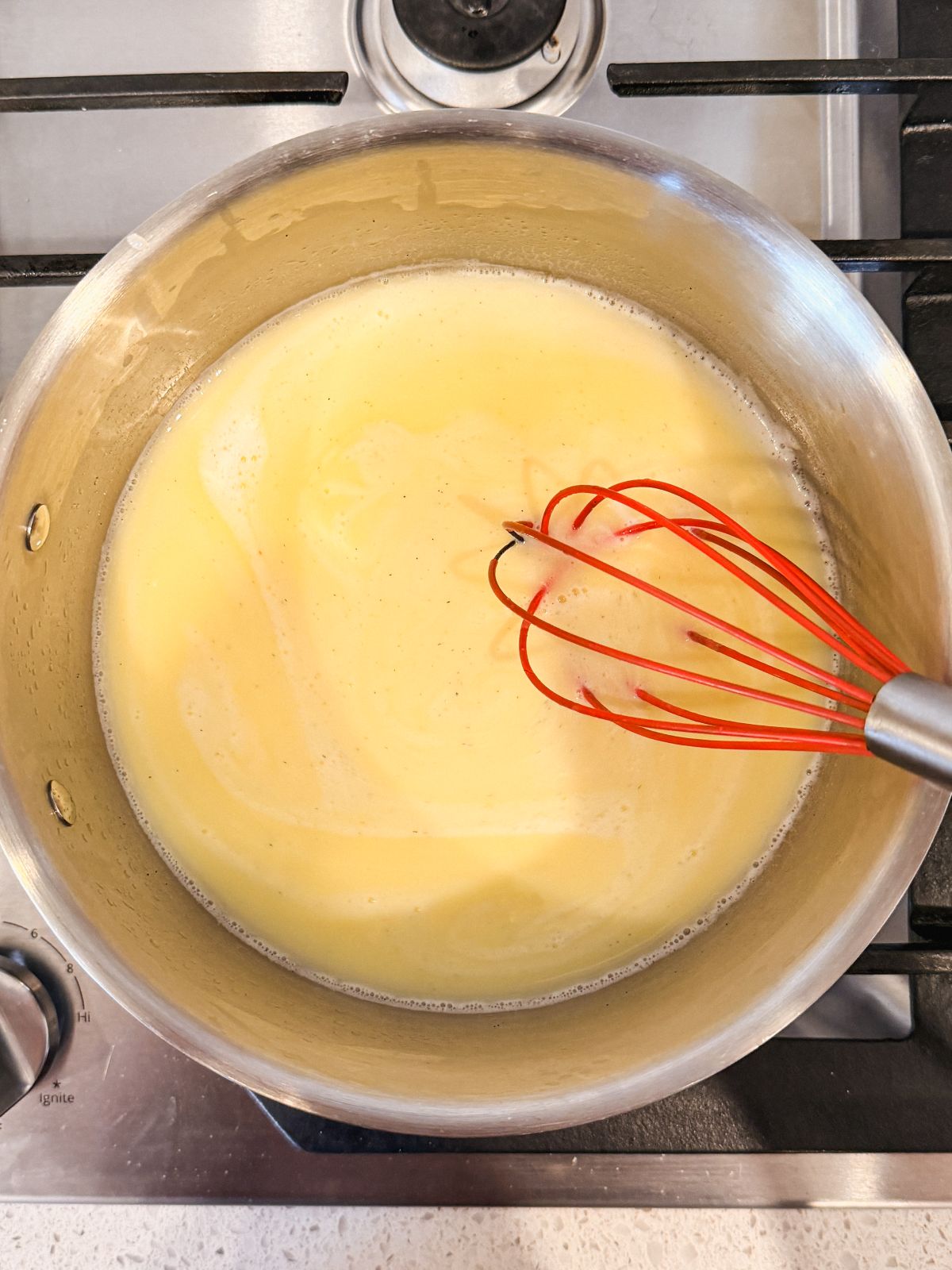 A red whisk stirring together a mixture of wet ingredients for choux dough in a stainless steel pot on a stove.