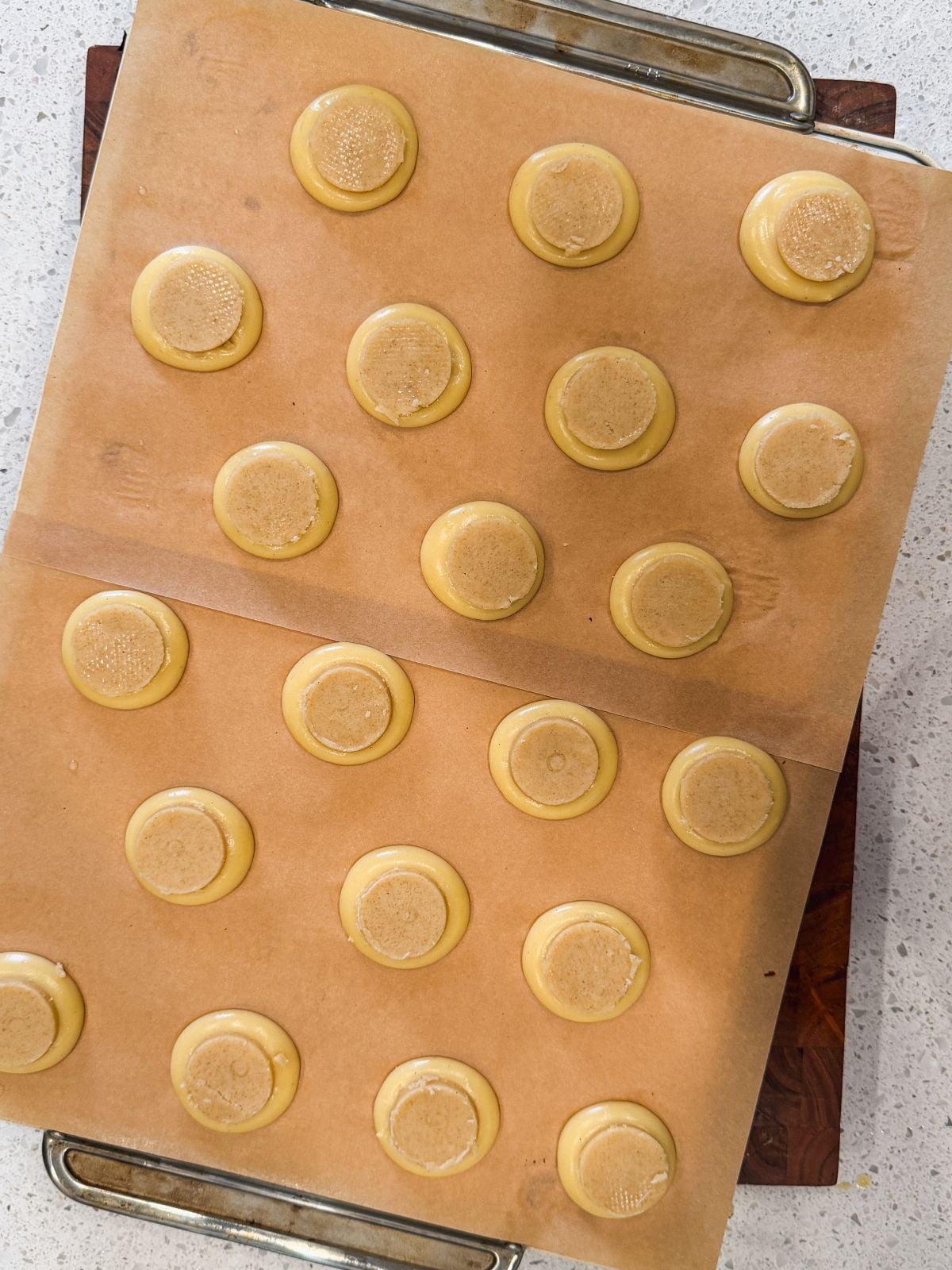A baking tray lined with parchment paper holds rows of piped choux pastry buns topped with craquelin circles, evenly spaced and ready to be baked. The tray is placed on a countertop.