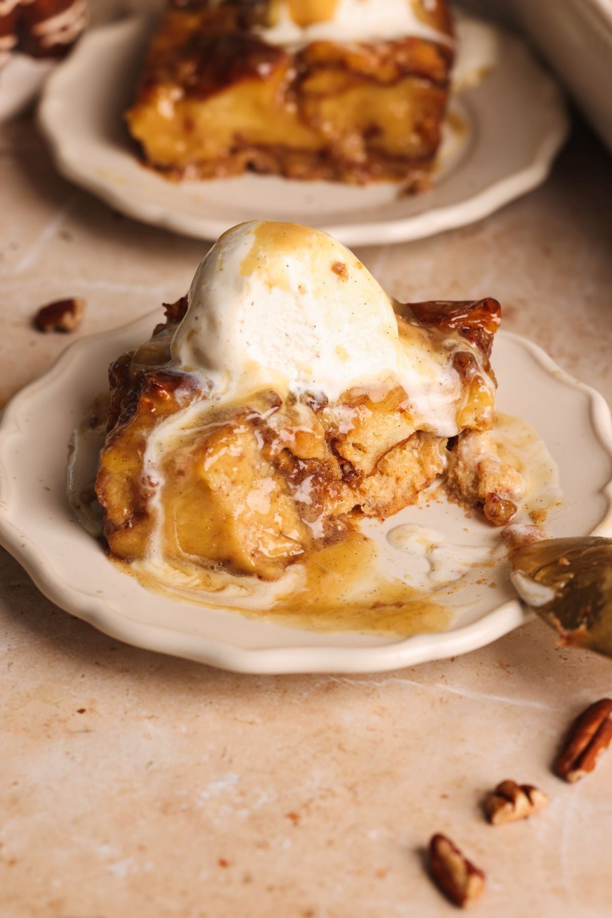 A slice of sticky toffee bread pudding topped with a scoop of vanilla ice cream and toffee sauce sits on a small plate, with a fork and pecan pieces nearby. The dessert appears gooey and partially eaten.