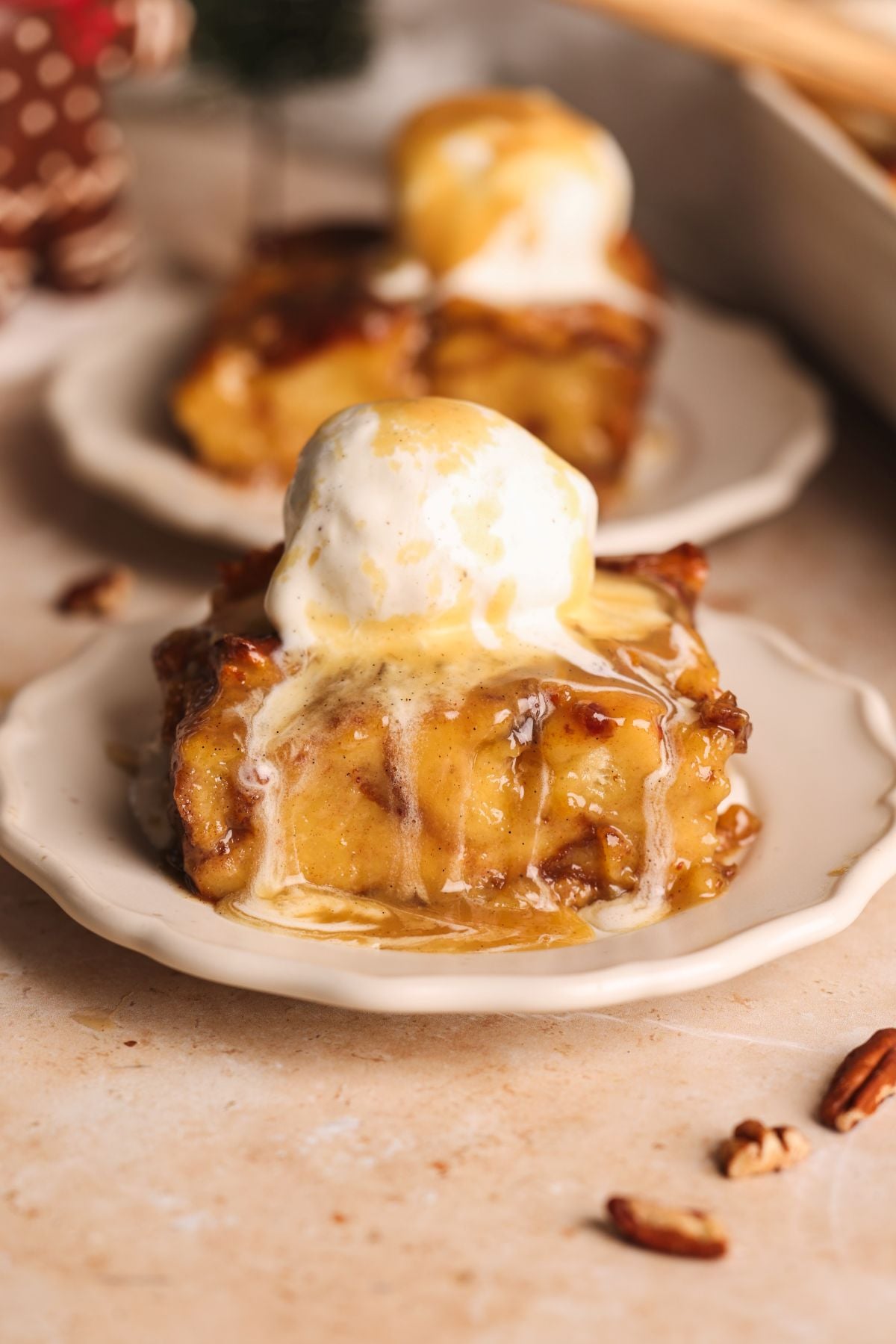 A slice of sticky toffee bread pudding topped with a melting scoop of vanilla ice cream sits on a small white plate. Another similar dessert is blurred in the background. Pieces of pecan are scattered nearby.
