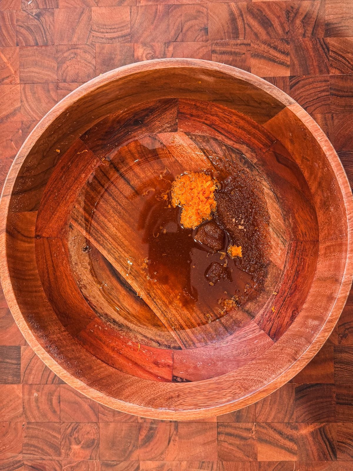 A large wooden bowl with small amounts of brown sugar, orange zest, and oil at the bottom, sitting on a wooden surface.