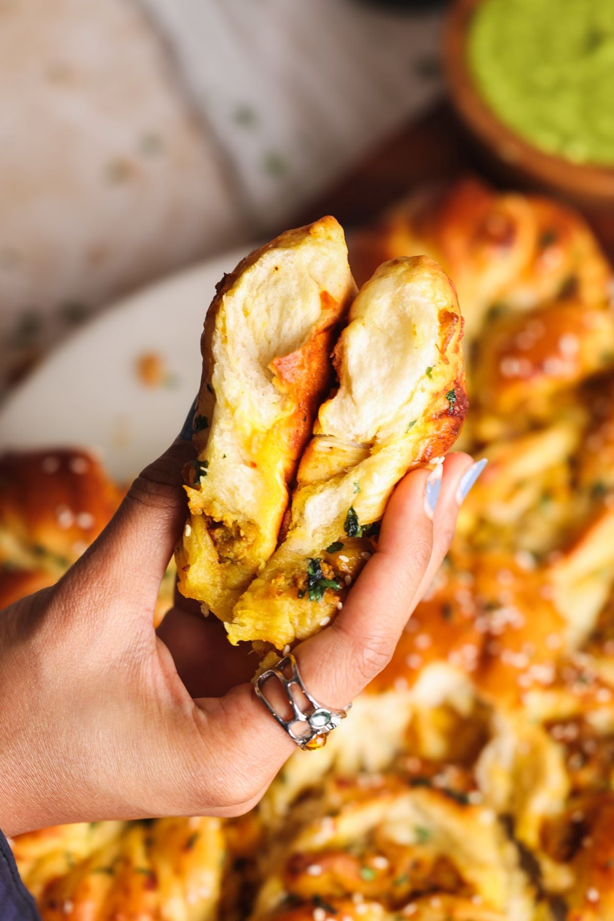 A hand holding and pulling apart a soft, golden, twisted bread filled with herbs and spices. The bread has a fluffy texture and visible green and orange filling. Other bread is blurred in the background.