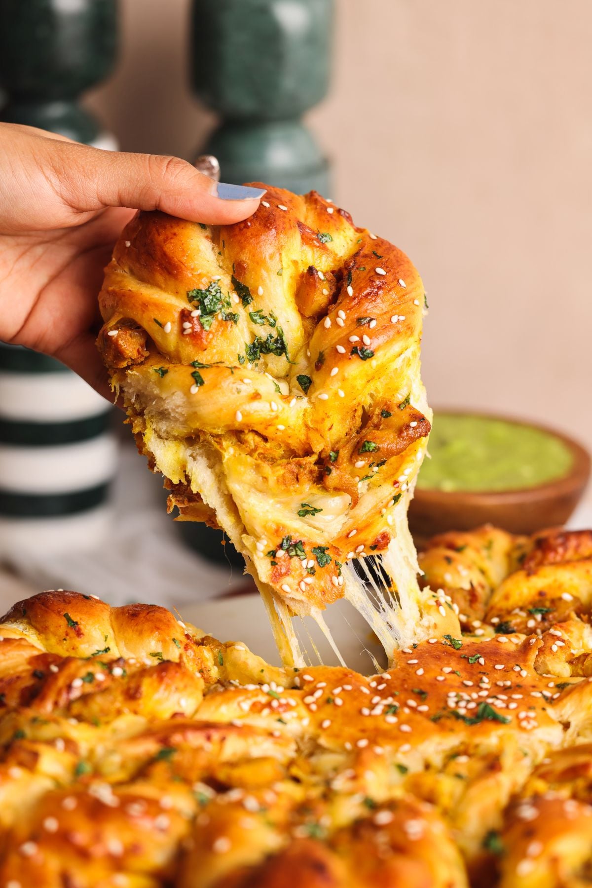 A hand pulls apart a piece of golden, cheesy tandoori chicken bread topped with herbs and sesame seeds, revealing gooey melted cheese inside. A bowl of green dipping sauce sits in the background.
