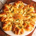 A round, golden-brown star-shaped bread with twisted segments filled with a herbed mixture, topped with sesame seeds and chopped parsley, served on a white plate.