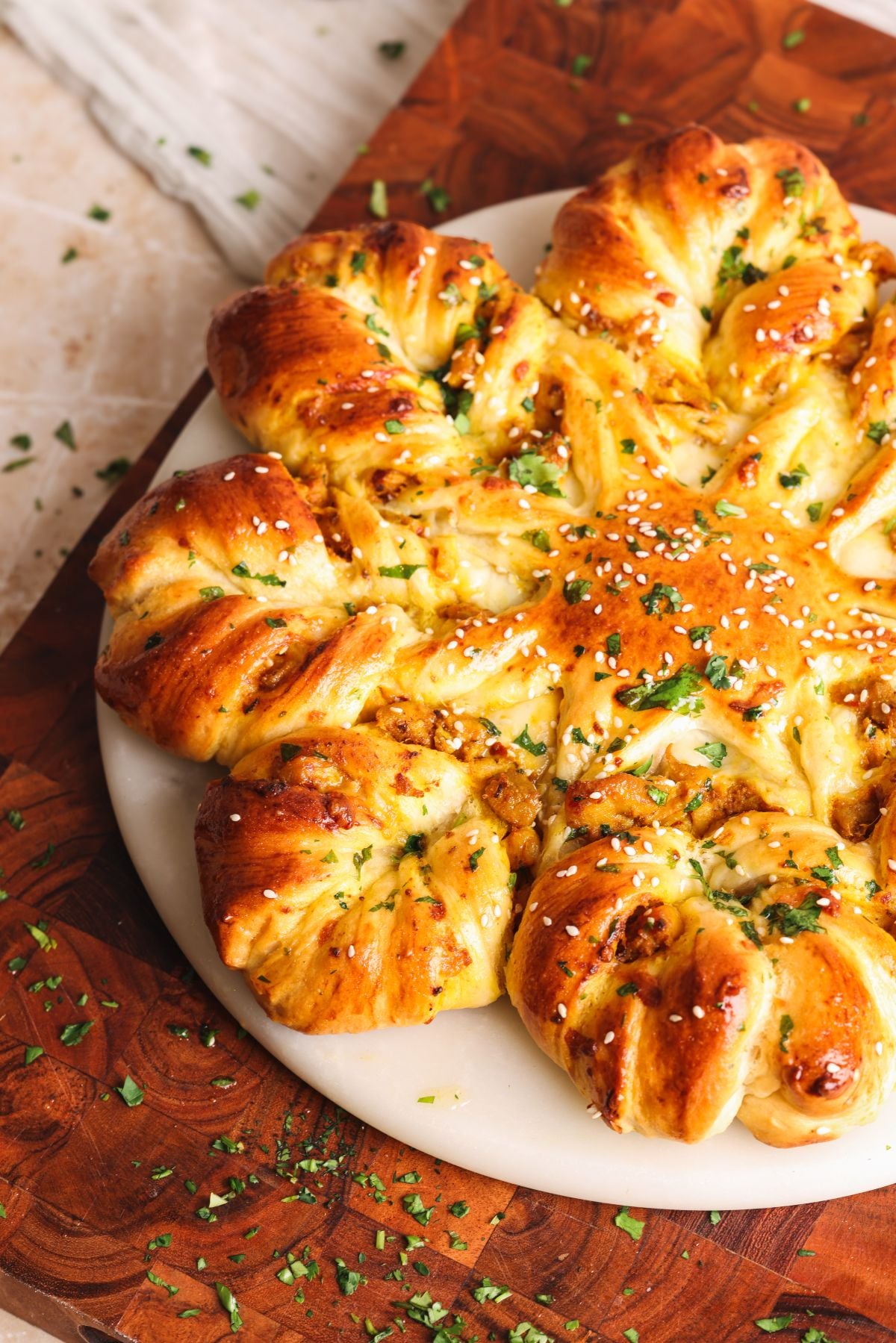 A round, golden-brown star bread with twisted segments, sprinkled with sesame seeds and chopped herbs, sits on a white plate atop a wooden board. Crumbs and herbs are scattered around.