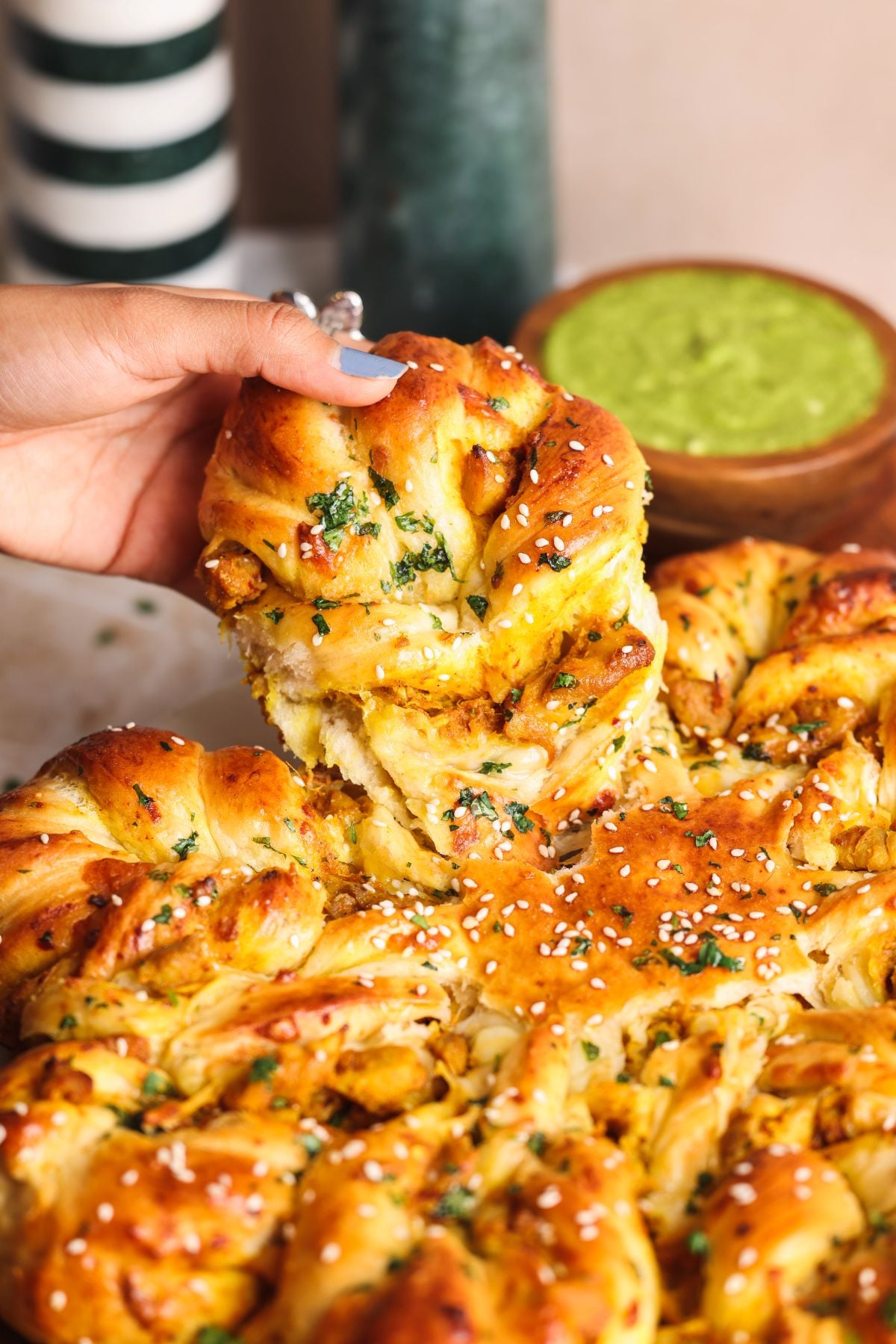 A hand pulls apart a piece of golden, twisted bread topped with herbs and sesame seeds. A bowl of green dip sits in the background. The bread appears soft and freshly baked.