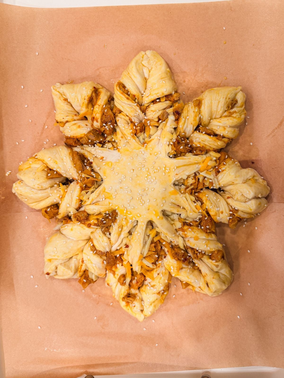 A star-shaped, twisted bread pastry filled with a savory mixture, topped with sesame seeds, sits on a sheet of brown parchment paper before baking.