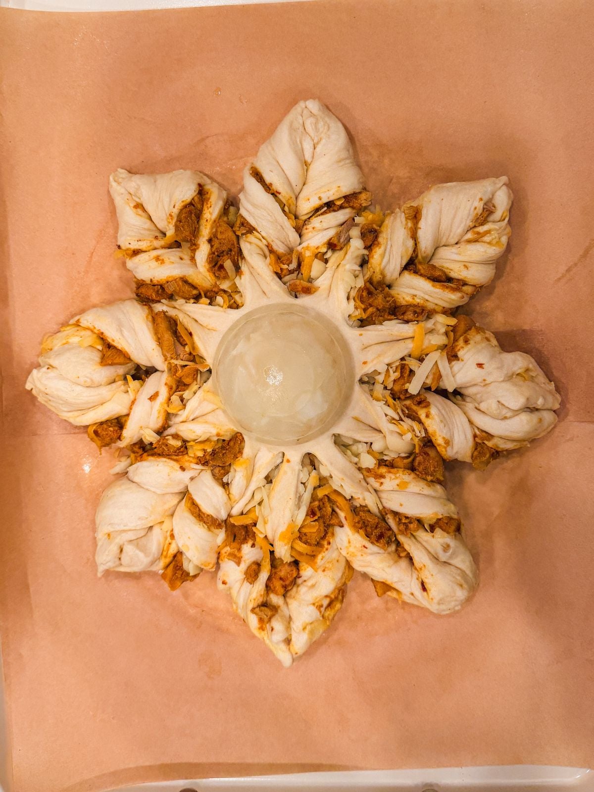 A star-shaped bread wreath with twisted, filled sections of dough arranged around a glass cup in the center, placed on parchment paper. The filling appears savory, with shredded meat and cheese.