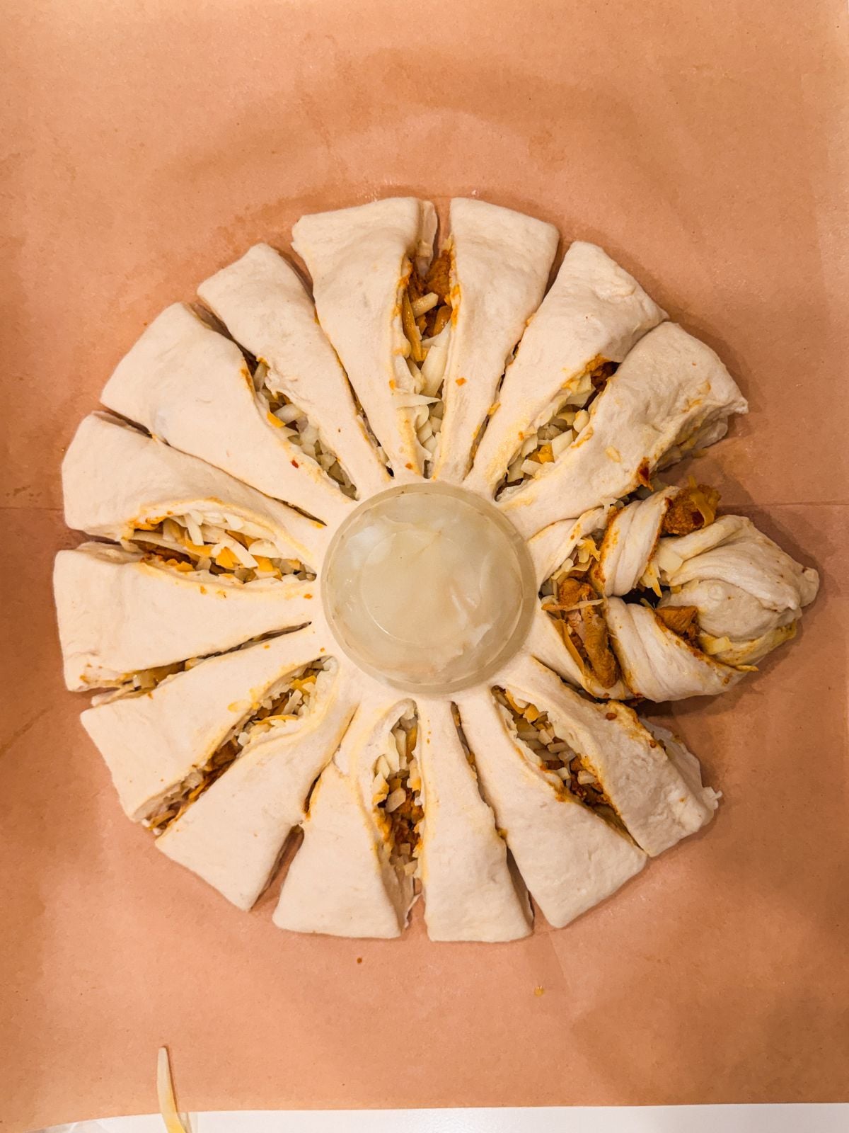 A round, flower-shaped arrangement of stuffed bread slices surrounds a small cup of white dipping sauce, all placed on brown parchment paper. The bread appears to have a savory filling.