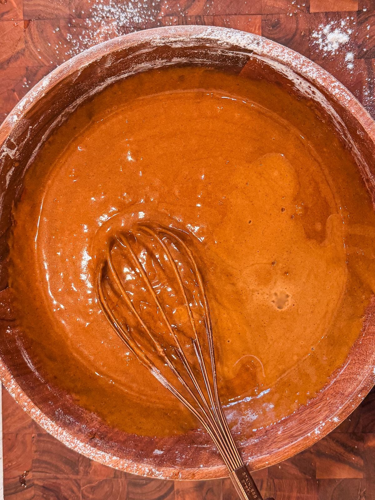 A metal whisk rests in a large wooden bowl filled with smooth, brown gingerbread cake batter on a wooden surface dusted with flour.