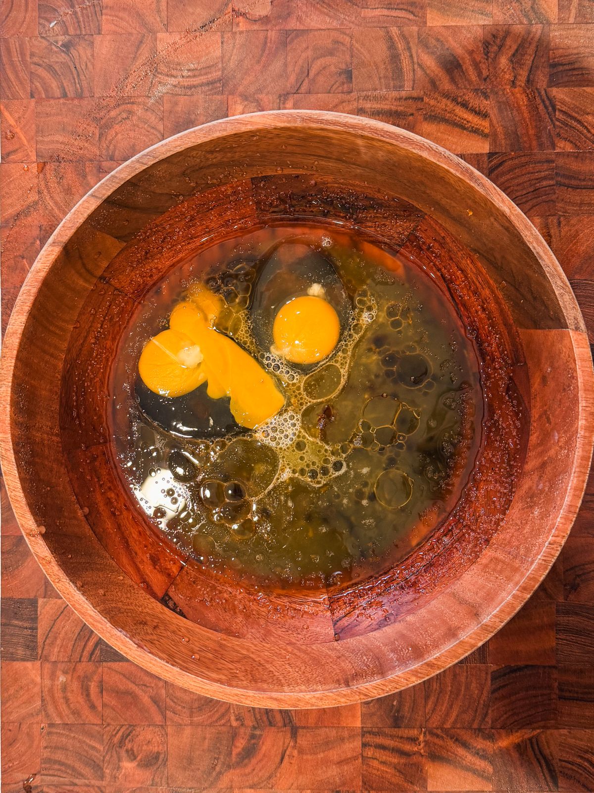 A wooden bowl on a wooden surface contains cracked raw eggs, with visible yolks and whites mixing together.