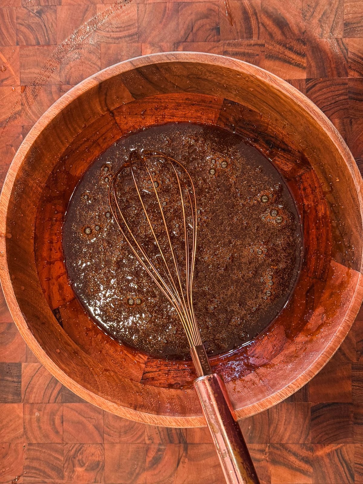 A metal whisk rests in a wooden bowl filled with a dark, liquid mixture on a wood-patterned surface. Bubbles and specks are visible in the mixture, suggesting it is being prepared for baking or cooking.