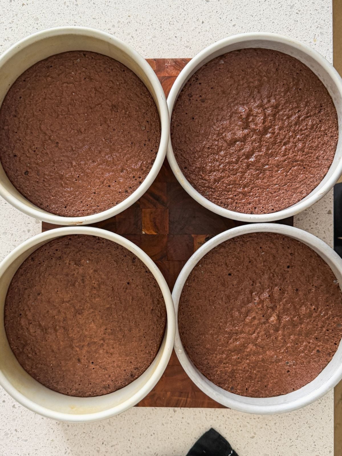 Four round cake pans filled with freshly baked chocolate cakes are arranged in a square on a wooden cutting board, set on a light-colored countertop.
