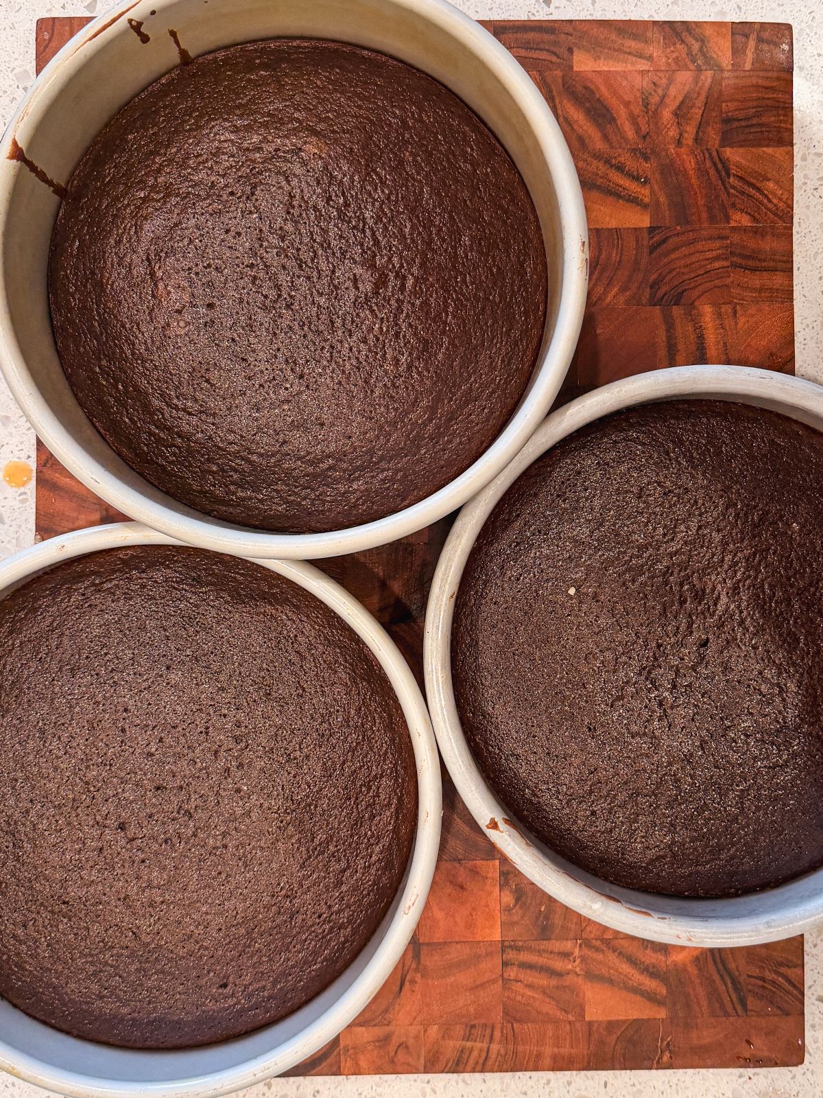 Three round pans filled with freshly baked chocolate cakes rest on a wooden cutting board, viewed from above. The cakes have smooth, dark tops and look evenly baked.