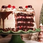 A rich, layered black forest cake with cherries, chocolate drizzle, and whipped cream sits on a green cake stand, with a slice removed to show the layers. Festive holiday decorations are in the background.