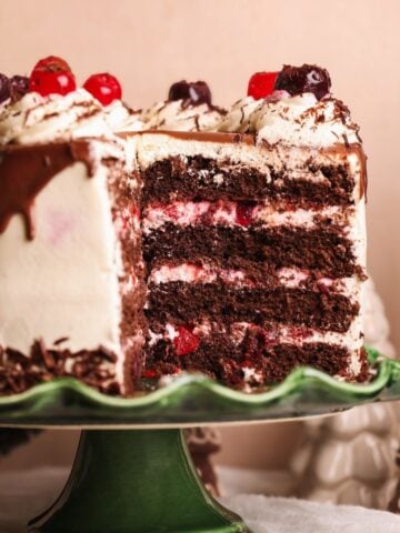 A rich, layered black forest cake with cherries, chocolate drizzle, and whipped cream sits on a green cake stand, with a slice removed to show the layers. Festive holiday decorations are in the background.
