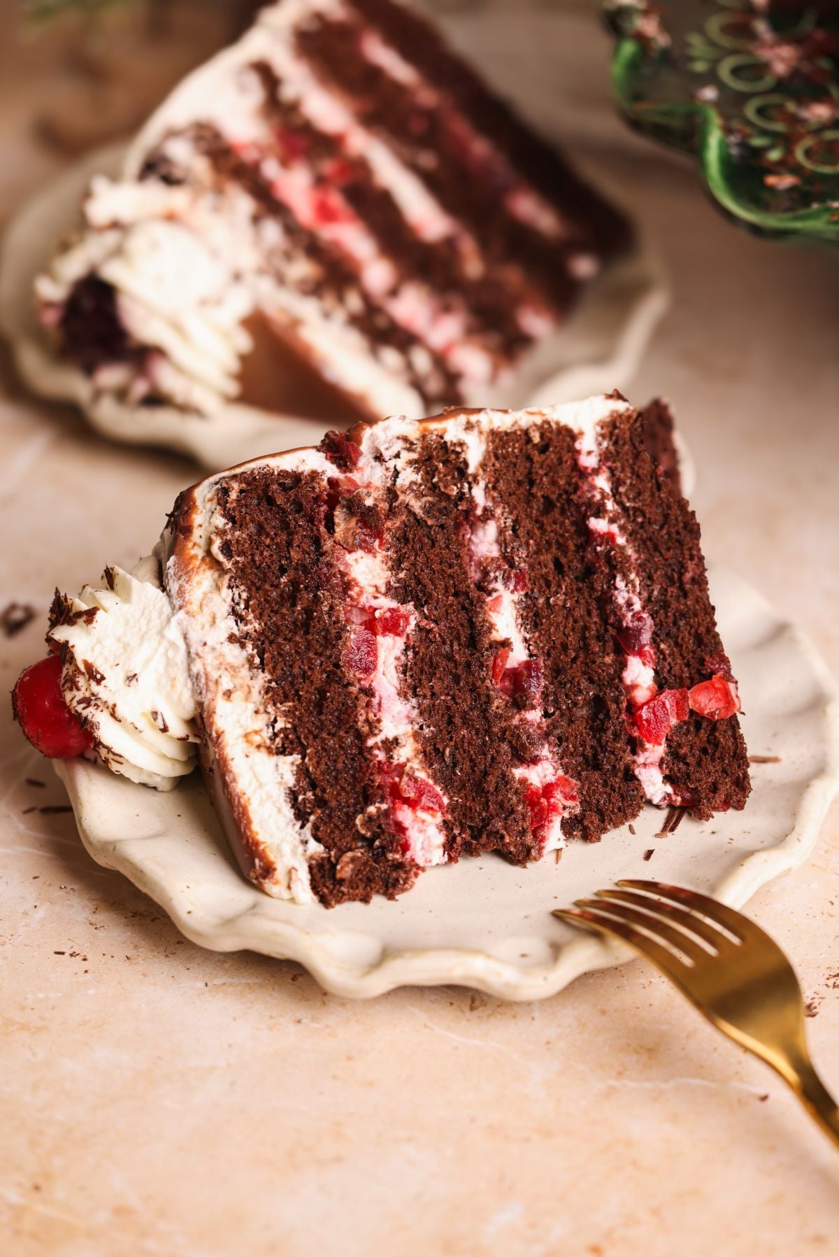 A slice of black forest cake with white cream and red cherries, served on a scalloped plate with a gold fork. Another slice of cake is blurred in the background.