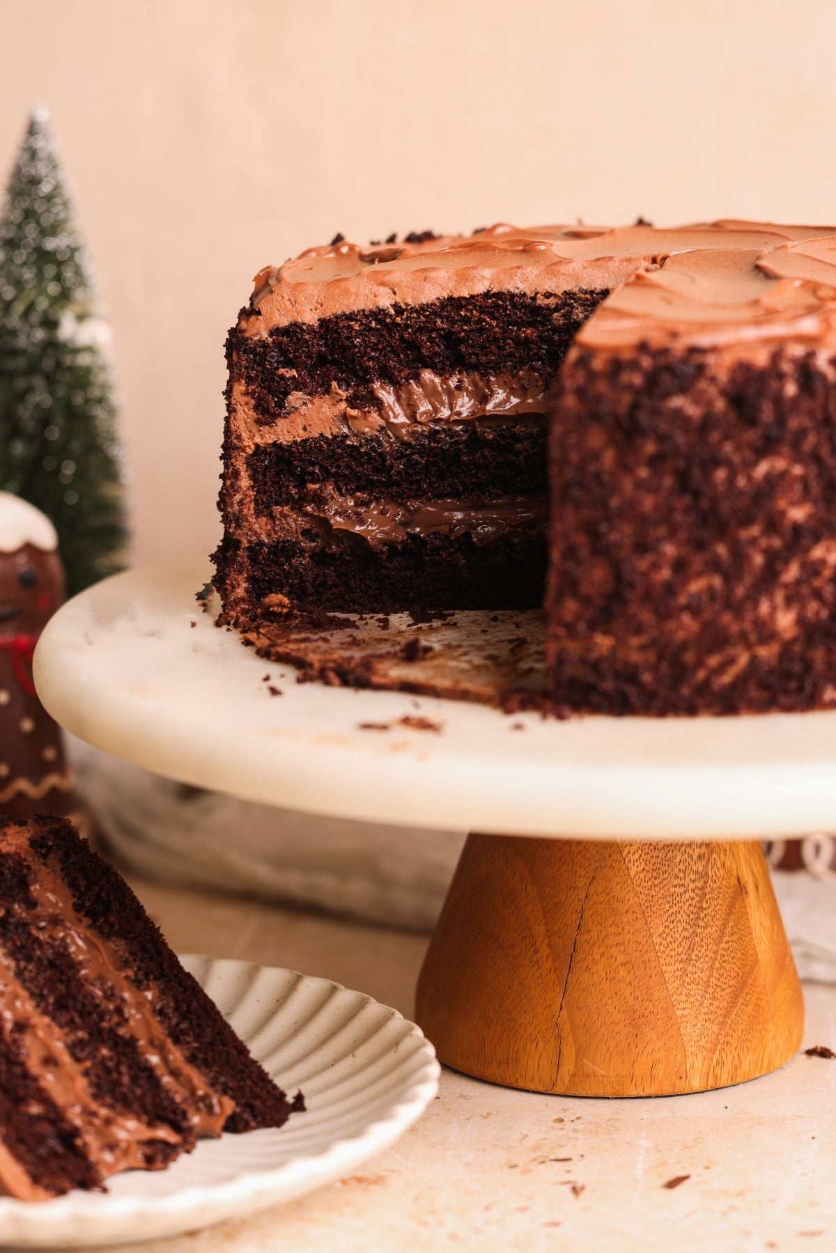 A three-layered chocolate cake with chocolate pudding filling sits on a cake stand. A large slice has been cut and placed on a white plate in the foreground. Small decorative trees are blurred in the background.