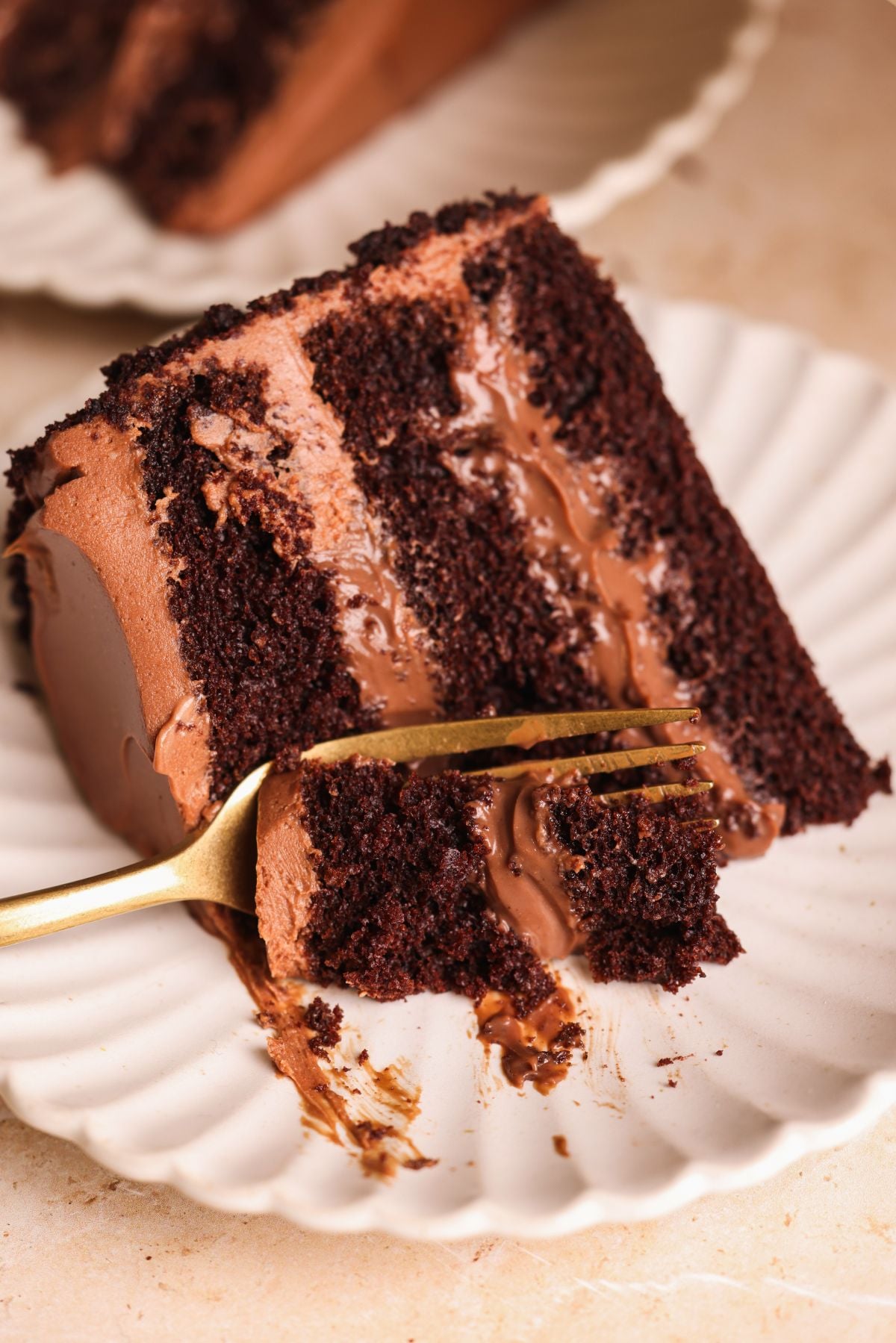 A close-up of a slice of rich brooklyn blackout chocolate cake with creamy chocolate pudding, served on a white scalloped plate. A gold fork holds a bite of the moist cake, showing its fluffy texture and layers.