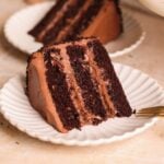 A slice of rich, three-layer chocolate cake with chocolate frosting and chocolate pudding filling sits on a white scalloped plate, with a fork nearby and another slice in the background.
