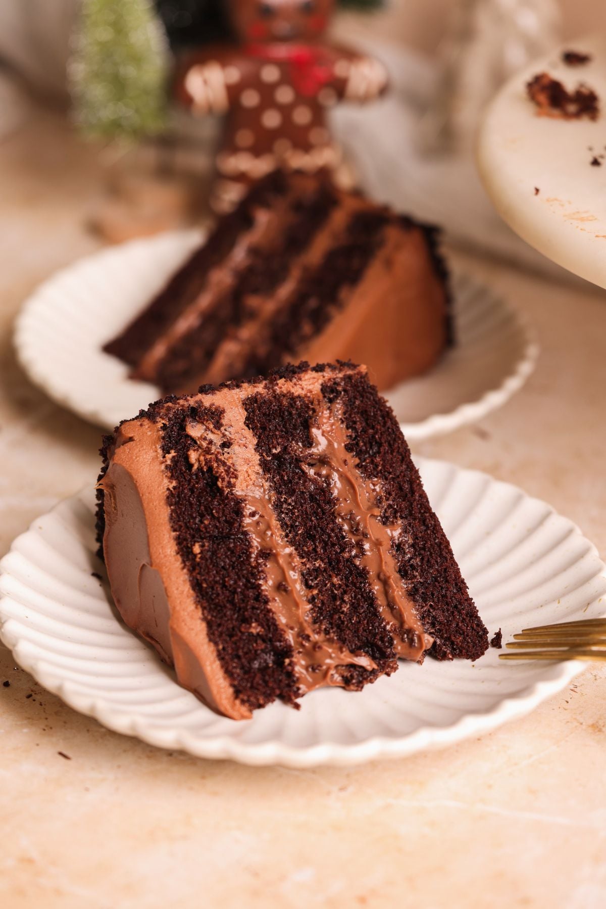 A close-up of two slices of rich chocolate layer cake with chocolate frosting on white plates, one in the foreground with a fork beside it, and festive holiday decorations blurred in the background.