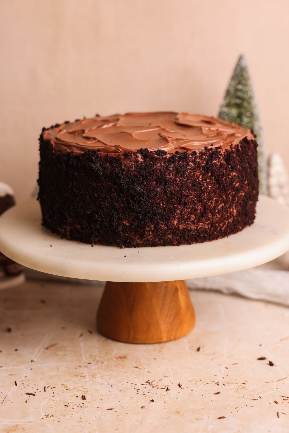 A round brooklyn blackout chocolate cake with chocolate frosting on top sits on a marble cake stand with a wooden base. The cake is decorated with chocolate crumbs around the sides, and there are blurred holiday decorations in the background.