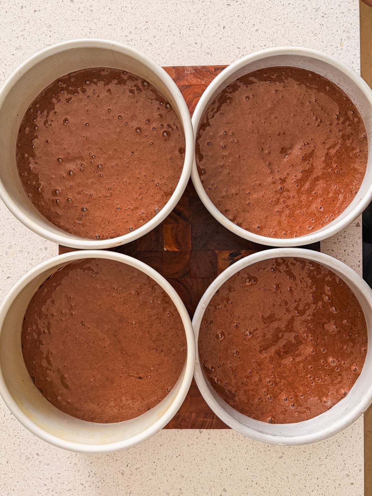 Four round baking pans filled with chocolate cake batter are arranged in a square on a countertop, ready to be baked.