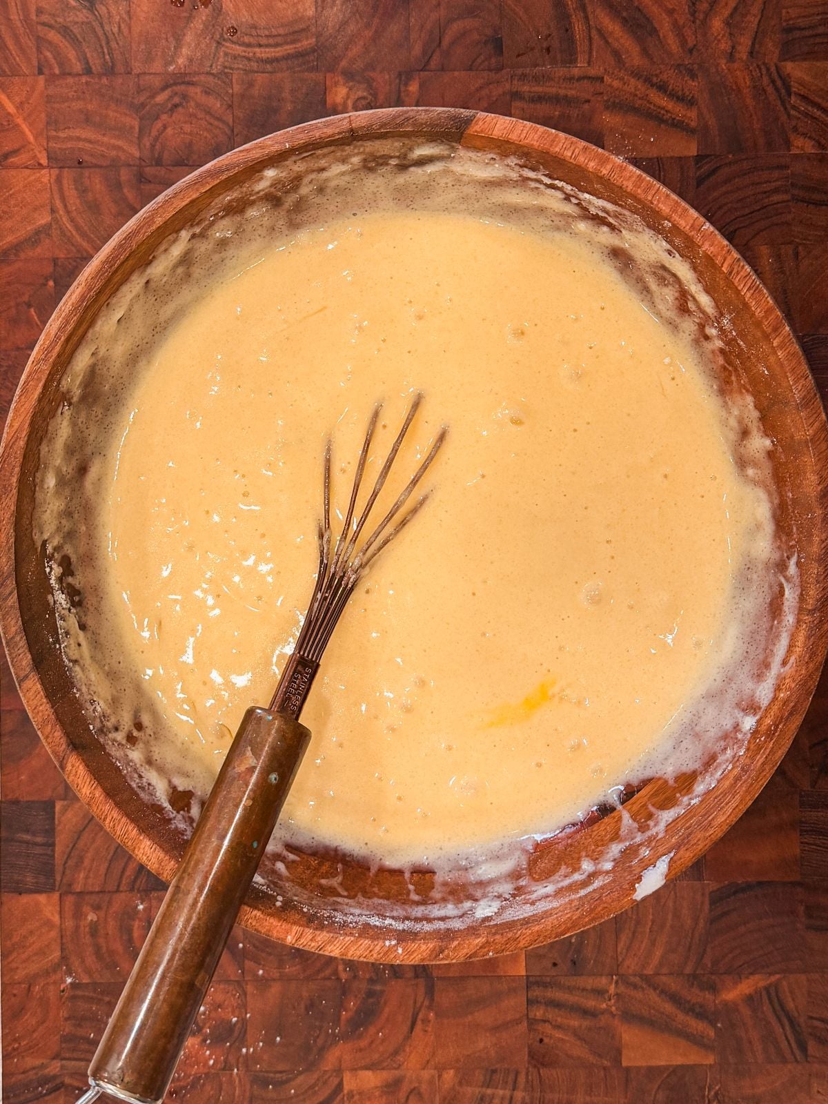 A wooden bowl filled with creamy, pale yellow cake batter sits on a wooden surface. A metal whisk with a brown handle rests inside the bowl, partially covered in the thick batter.